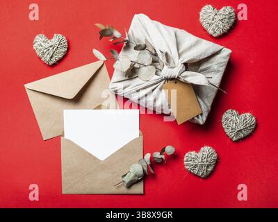 Zero waste Valentine's Day concept and mock up on red. Eco-friendly gift cloth wrapping in Furoshiki style, craft paper envelope, empty greetings card Stock Photo