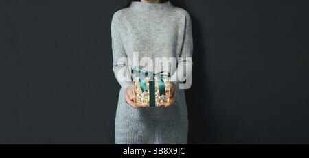 Woman in gray pullover holding luminous gift box in hands on dark background. Glowing yellow light bulbs into transparent gift box with dark green rib Stock Photo