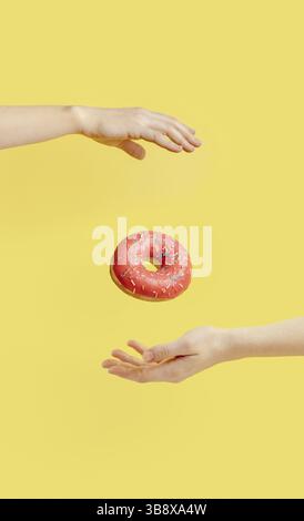 Donut falls between hands on bright yellow paper background. Donuts delivery, diet, sugar concept. Female hands and glazed donut with sprinkles flying Stock Photo