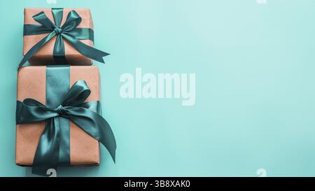 Two gift boxes in craft wrapping paper and green satin ribbon on turquoise blue background, copy space right. Beautiful Christmas, New Year or Birthda Stock Photo