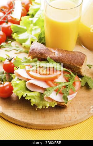 Wholegrain sandwich with ham, tomato, lattuce and arugula with glass of orange juice on the board Stock Photo