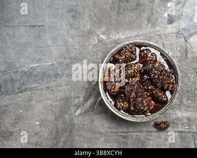 Delicious jam with baby pine cones in small bowl. Traditional Siberian dessert - young pine cones jam on gray textured background. Top view or flat la Stock Photo
