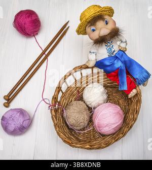 From above the colored balls of yarn in wicker basket with needles and traditional toy of man on white wooden board in Ukraine Stock Photo
