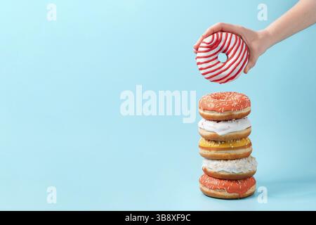 Stack of assorted donuts and one donut in female hand on blue background. Many colorful glazed doughnut with sprinkles stacked with copy space Stock Photo