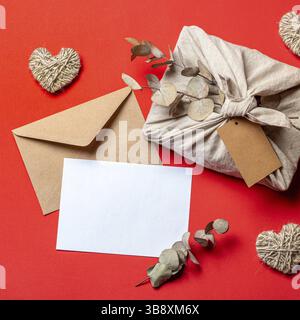 Zero waste Valentine's Day concept and mock up on red. Eco-friendly gift cloth wrapping in Furoshiki style, craft paper envelope, empty greetings card Stock Photo