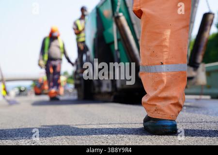 Low Angle Shot of Construction Worker’s Leg on Road Renovation Site with Blurred Equipment and Workers in the Background Stock Photo