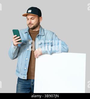 Casual man in snapback leaning on blank placard while using smartphone. Stock Photo