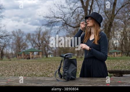 A young woman stands outdoors in a park, applying lipstick, holding a small mirror. Stock Photo
