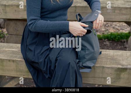 A girl wearing a dark blue outfit sits on a wooden bench, opening a gray backpack. Stock Photo