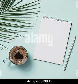 Blank paper notebook with pencil and coffee cup. Empty paper sketchbook with pincil and tropical leaves on blue background. Top view or flat lay. Copy Stock Photo