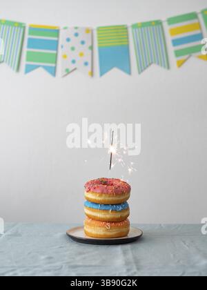 Birthday donuts cake with bengal light or sparkler. Stack of delicious doughnuts over table with party flag garland on background. Vertical. Copy spac Stock Photo