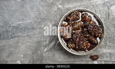 Delicious jam with baby pine cones in small bowl. Traditional Siberian dessert - young pine cones jam on gray textured background. Top view or flat la Stock Photo