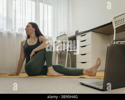 Young woman sitting on floor and doing yoga or stretching at home near laptop. Yoga online in small space home and wellbeing concept. Copy space for t Stock Photo