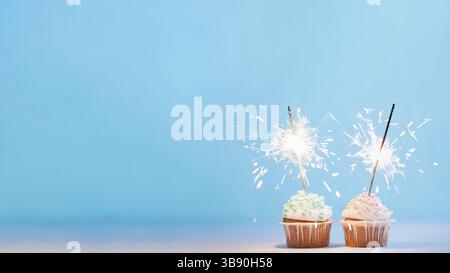 Two cupcakes decorated with colorful sprinkles and sparklers. Festive cupcakes on blue background with copy space left for text or design. Banner Stock Photo