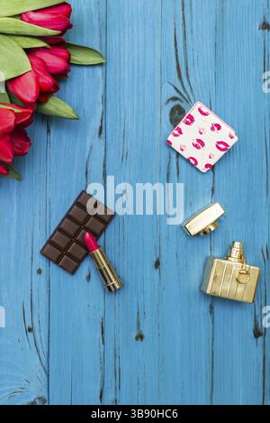 Red tulips in the necklace, lipstick, bottle of perfume and piece of chocolate on the background of blue wooden board Stock Photo