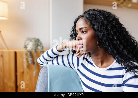 Sad young African American woman sitting on sofa looking away at home Stock Photo