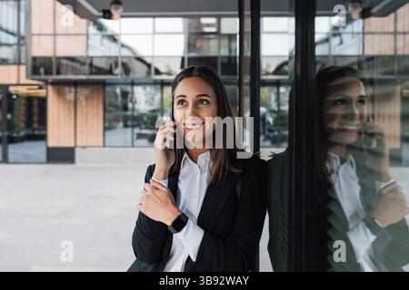 Smiling female in black formal wear leaning business building and talking on mobile phone. Female office worker talking on mobile phone during a break Stock Photo