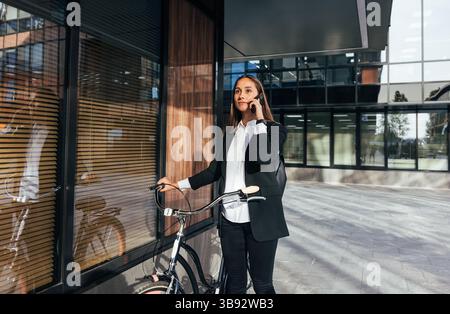 Young female entrepreneur talking on mobile phone while walking outdoors with bicycle. Woman in formal wear is talking on mobile phone while standing Stock Photo
