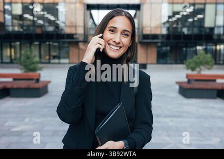 Portrait of a smiling woman employee in black formal wear. Happy businesswoman wearing stylish formal wear, talking on mobile phone and looking at cam Stock Photo
