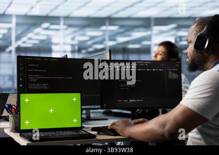 Isolated screen laptop next to man coding in server farm while listening music in headphones. Worker safeguarding hardware against unauthorized access with chroma key notebook, hearing songs Stock Photo
