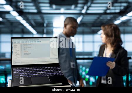 Close up of code lines on laptop screen in front of data center engineers checking documents. Focus on programming language on notebook display next to team of server farm admins reading paperwork Stock Photo