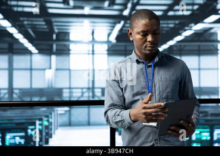 Computer scientist in data center uses tablet, verifying compliance of racks. IT expert in high tech facility optimizing resource utilization, maintaining stable infrastructure using device Stock Photo