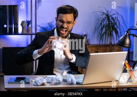 Angry furious Indian man working at office throwing crumpled paper, having nervous breakdown at work, migraine headache, stress management, mental distress problems, losing temper, reaction on failure Stock Photo