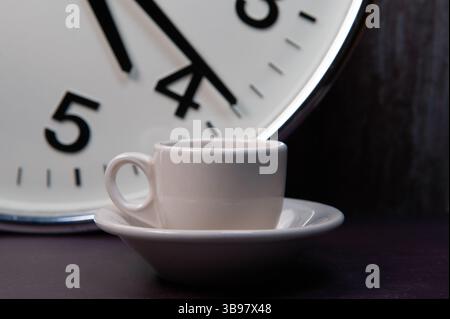 A white ceramic cappuccino cup and saucer in front of a large industrial clock. Stock Photo