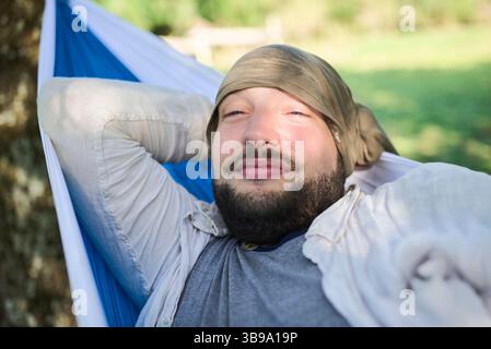 Young sleepy man waking up from nap, resting under the shade of trees in a hammock hanging outdoors. Concept: enjoy vacations in contact with nature. Stock Photo