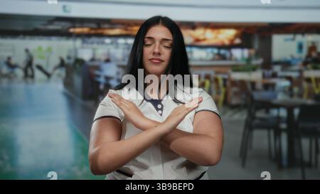 Woman with crossed arms in an indoor restaurant setting, wearing a white uniform, appears confident in a bustling coffee shop environment. Stock Photo