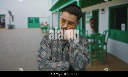 Young man with sunglasses relaxes on an outdoor restaurant terrace, wearing a patterned shirt, displaying a pensive expression amidst green decor and Stock Photo