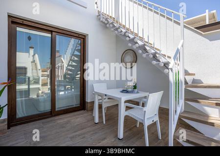 Charming lower-level terrace with a white table and chairs, string lights on staircase, and a cozy wine and snack setup. Stock Photo