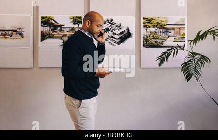 A professional architect talks on the phone while walking near displayed architectural designs in a creative office space. Modern architectural planni Stock Photo