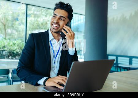 A professional Indian businessman in a corporate office engaging in a conversation on his smartphone while using a laptop. He appears confident and fo Stock Photo