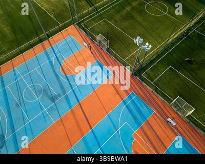 Outdoor basketball and soccer court at sunset with colorful surface, long shadows, and green artificial turf in the background Stock Photo