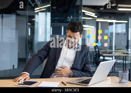 Man at workplace suffering from stomach pain. Businessman in business suit sitting at desk with laptop, patient holding stomach. Stock Photo