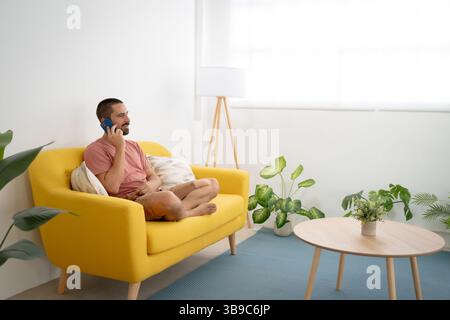 Smiling young man talking smartphone sitting on sofa at home Stock Photo