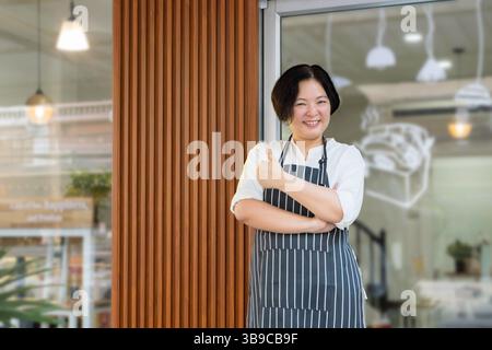 successful small business owner concept. a confident Asian woman wearing an apron, standing with her thumbs up with smiling face front of bakery shop Stock Photo