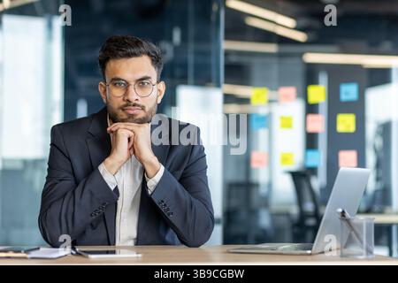 A thoughtful businessman, with glasses and beard, sitting in front of a laptop in a modern office. The man is wearing a suit and looking directly at the camera Stock Photo