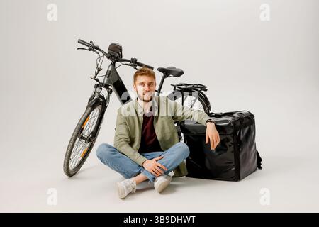 Young man sits on the floor next to his bicycle and delivery bag in a studio setting while preparing for work as a courier Stock Photo