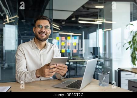A smiling programmer holding a tablet sits at a desk with a laptop. He looks happy and content in his modern office setting Stock Photo