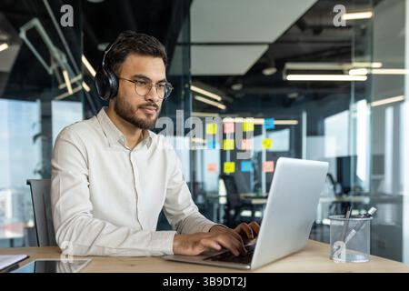 Serious thinking programmer coding new software. Man in headphones working with laptop inside office. Developer typing on keyboard. Stock Photo