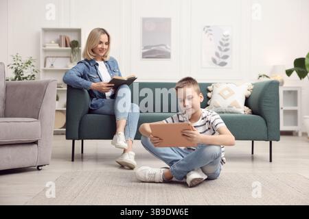 Son using tablet while his mother reading book at home, selective focus Stock Photo