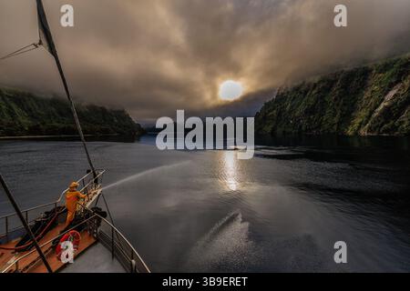 dramatic scene on doubtful sound cruise as yellow firefighter on prow sprays from high pressure hose towards sun rising behind stormy couds Stock Photo