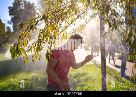 A happy young man stands in a sunny park, smiling with eyes closed as he enjoys the refreshing water spray from active sprinklers. His expression show Stock Photo