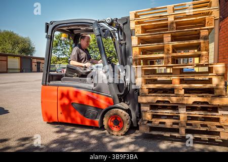 A man is driving a forklift with a stack of wood pallets on it. The forklift is red and black Stock Photo