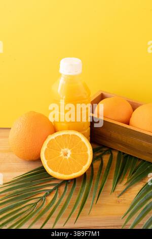 Orange juice bottle with fresh citrus fruits and palm leaf on wooden board with vibrant yellow background Stock Photo