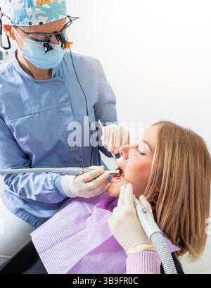 Dentist wearing magnifying glasses operating on patient's teeth using dental drill and saliva ejector in dental clinic Stock Photo