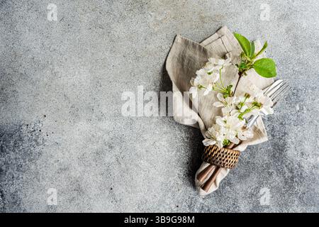 An elegant table setting with a folded linen napkin, delicate white flowers, and silver fork and knife on a textured stone surface creates a sophistic Stock Photo