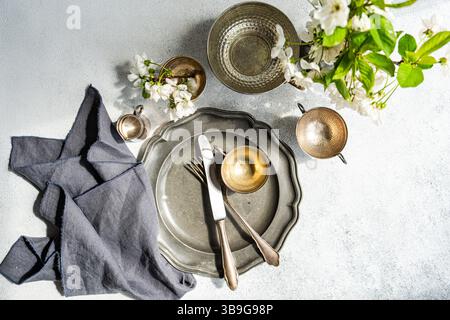 A top-down view of a table set with metal dishes, cutlery, and a gray linen napkin. Rustic elegance enhanced by delicate white blossoms in the decor. Stock Photo